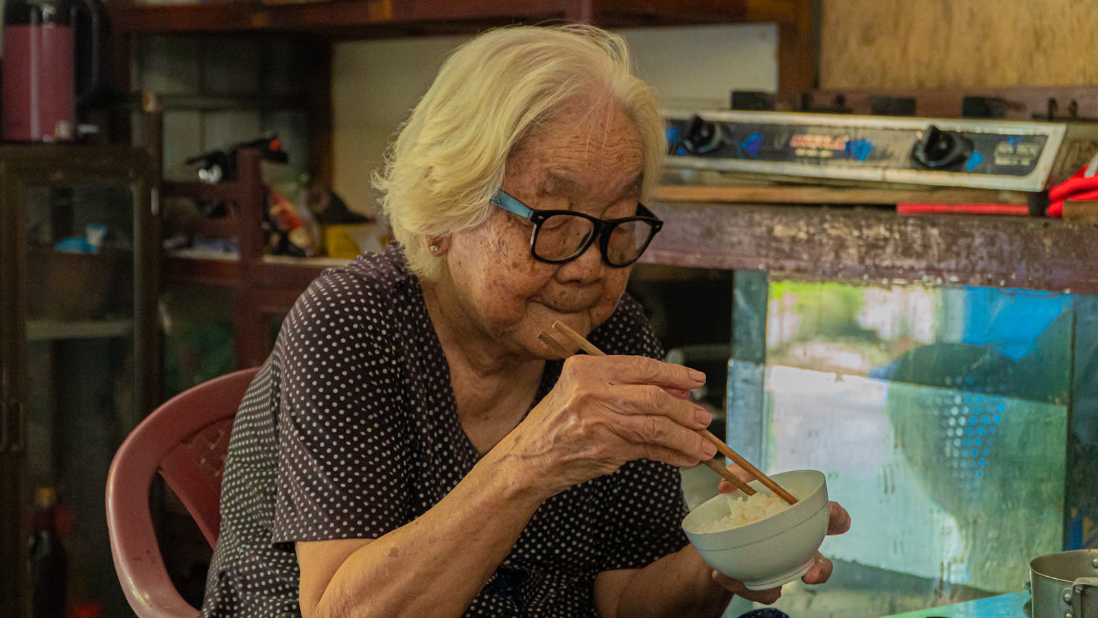 An older woman eating a bowl of rice with chopsticks at her kitchen table