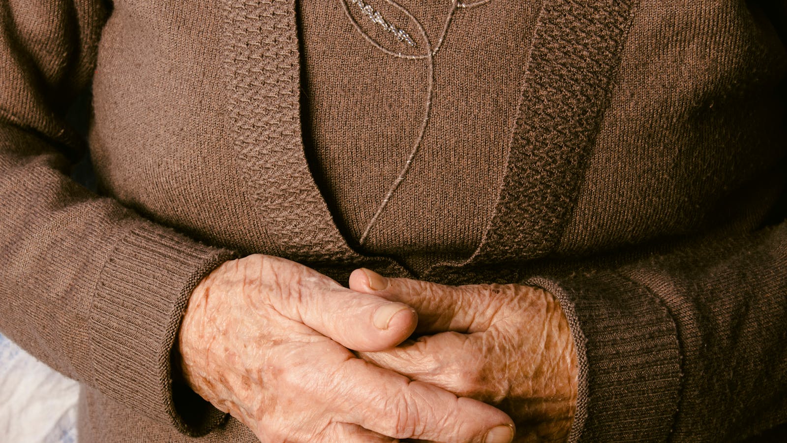 An older person's hands resting clasped over a brown knit cardigan